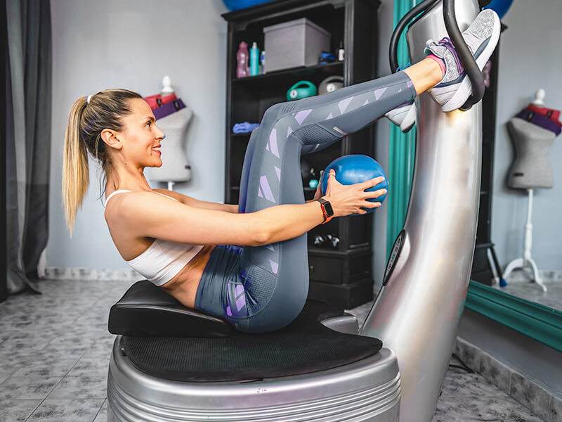 Patientin sitzend auf dem Power Plate Gerät. In den Händen hält Sie dabei einen kleinen Gymnastikball.