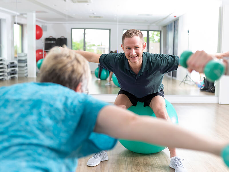 Therapeut und mehrere Patienten bei einer Ăbung mit Kurz-Hanteln und GymnastikbĂ€llen.