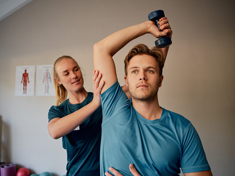 Physiotherapeutin hilft einem erwachsenen Patienten bei einer Ăbung mit einer kleinen Kurz-Hantel
